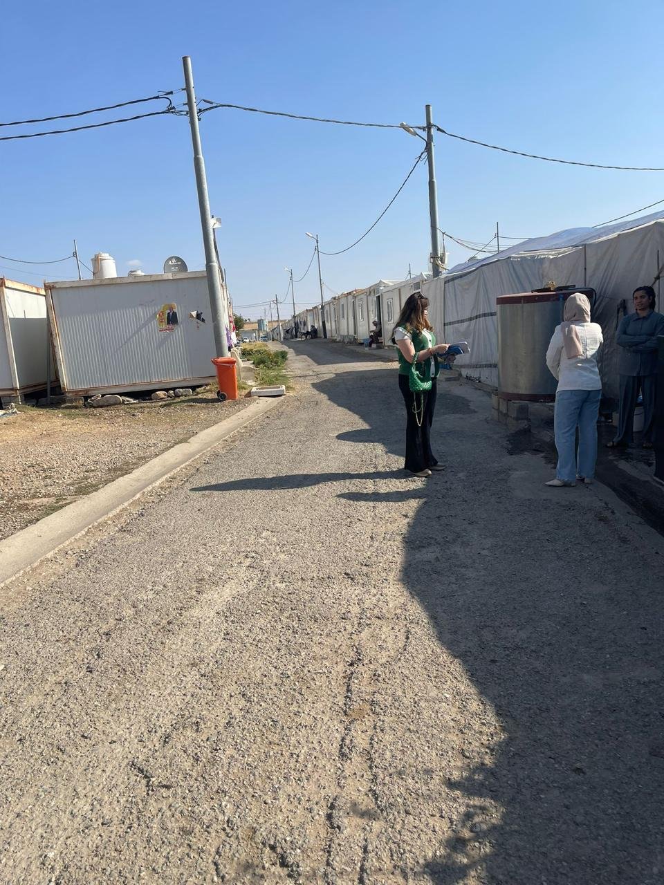 A KIANO staff member carrying assessment forms speaks with residents on a camp street lined with shelters