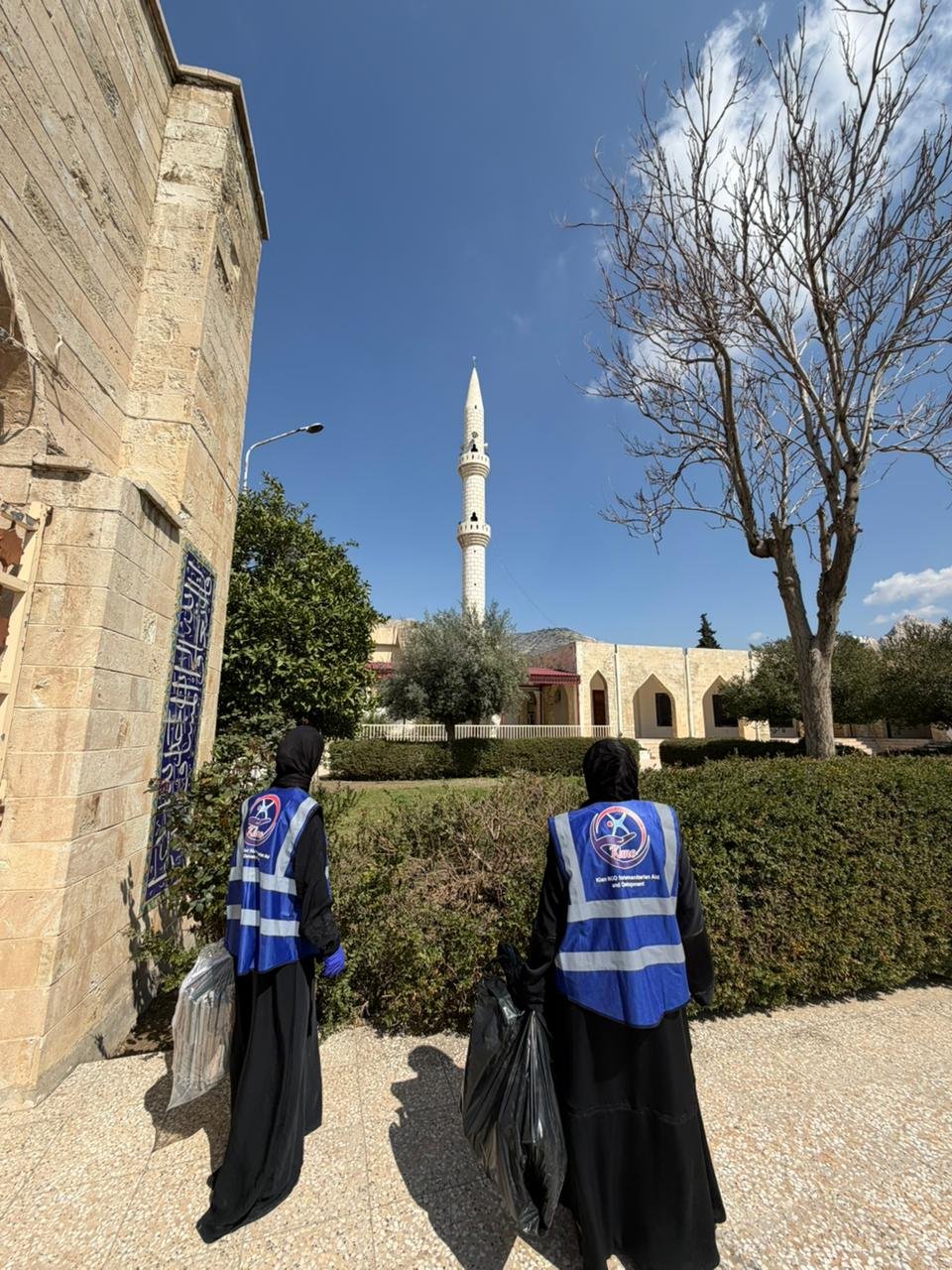 Two KIANO volunteers in blue vests carrying cleaning supplies towards a mosque courtyard in Duhok
